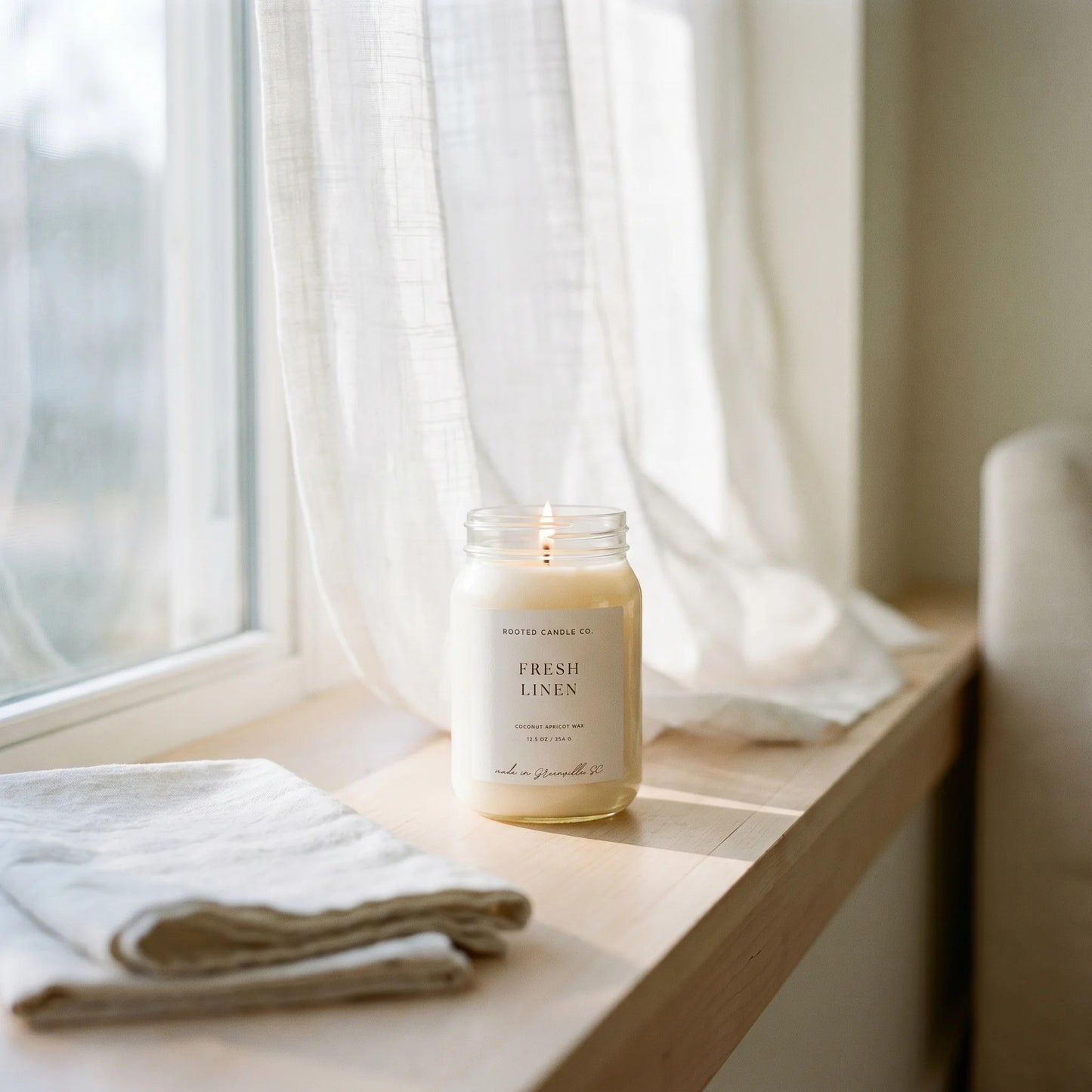 Fresh Linen candle on sunlit windowsill with billowing sheer curtain